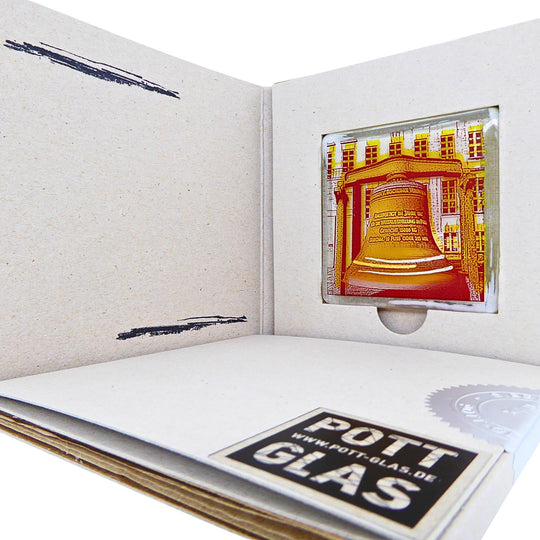 Bochum Edition 12/12. Geöffnete POTT GLAS Blanko-Inlay-Karte mit Beschriftungsfeld. Rechts ein Magnet-POTTRÄT aus Schmelzglas mit gelbem Rathausglocke-Motiv sowie Akzenten aus 24-Karat-Flüssiggold und Platin im Karten-Inlay mit Entnahmeaussparung. Darunter die versiegelte Karte mit Logo und geprägter Siegelplombe.