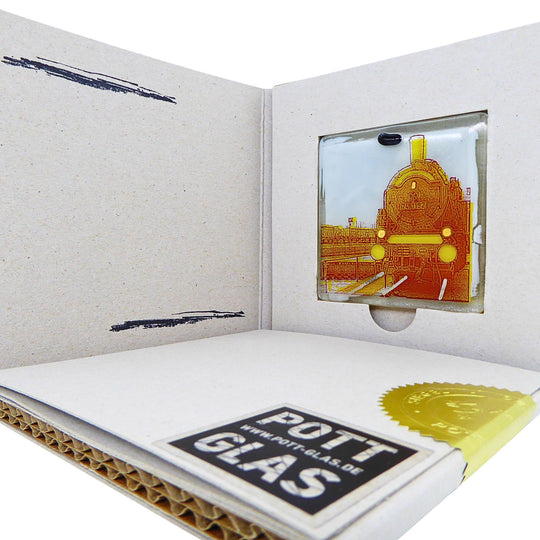 Bochum Edition 05/12. Geöffnete POTT GLAS Blanko-Inlay-Karte mit Beschriftungsfeld. Rechts ein Magnet-POTTRÄT aus Schmelzglas mit gelbem Dampflok-Motiv sowie Akzenten aus 24-Karat-Flüssiggold und Platin im Karten-Inlay mit Entnahmeaussparung. Darunter die versiegelte Karte mit Logo und geprägter Siegelplombe.