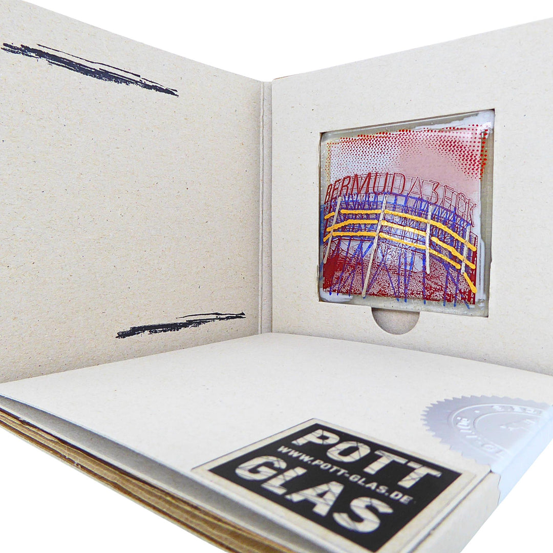Bochum Edition 06/12. Geöffnete POTT GLAS Blanko-Inlay-Karte mit Beschriftungsfeld. Rechts ein Magnet-POTTRÄT aus Schmelzglas mit blau-rotem Bermudadreieck-Motiv sowie Akzenten aus 24-Karat-Flüssiggold und Platin im Karten-Inlay mit Entnahmeaussparung. Darunter die versiegelte Karte mit Logo und geprägter Siegelplombe.